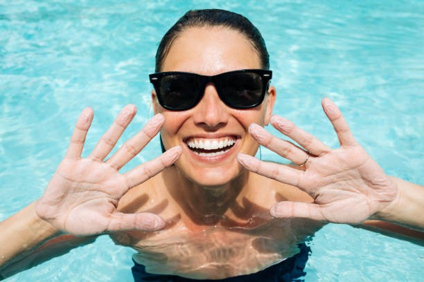 Smiling woman in the pool with sunglasses, showing off her pruny hands